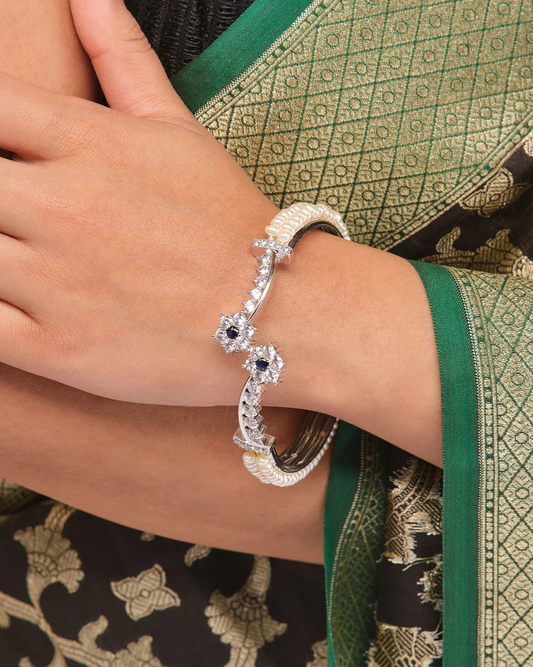 The Chandrani Pearls Floral Blossom Pearl CZ Bangle features small pearls, two star-shaped accents with blue and clear stones, and delicate floral CZ motifs, elegantly displayed on brown fabric.