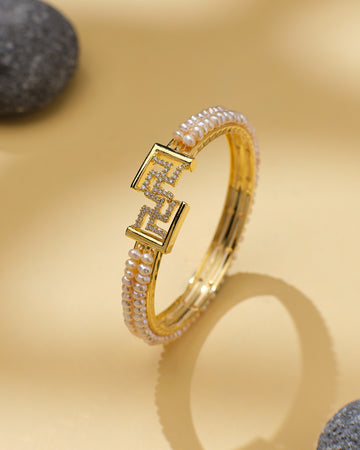 The Swasthik Pink Pearl & CZ Cuff Bangle by Chandrani Pearls is a chic statement piece, featuring sparkling CZ stones and pearls with a geometric clasp, displayed on beige beside gray stones.