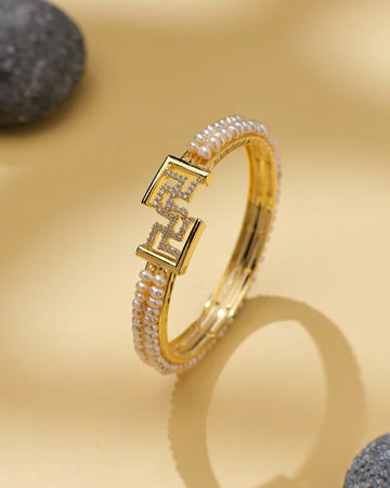 The Swasthik Pink Pearl & CZ Cuff Bangle by Chandrani Pearls is a chic statement piece, featuring sparkling CZ stones and pearls with a geometric clasp, displayed on beige beside gray stones.