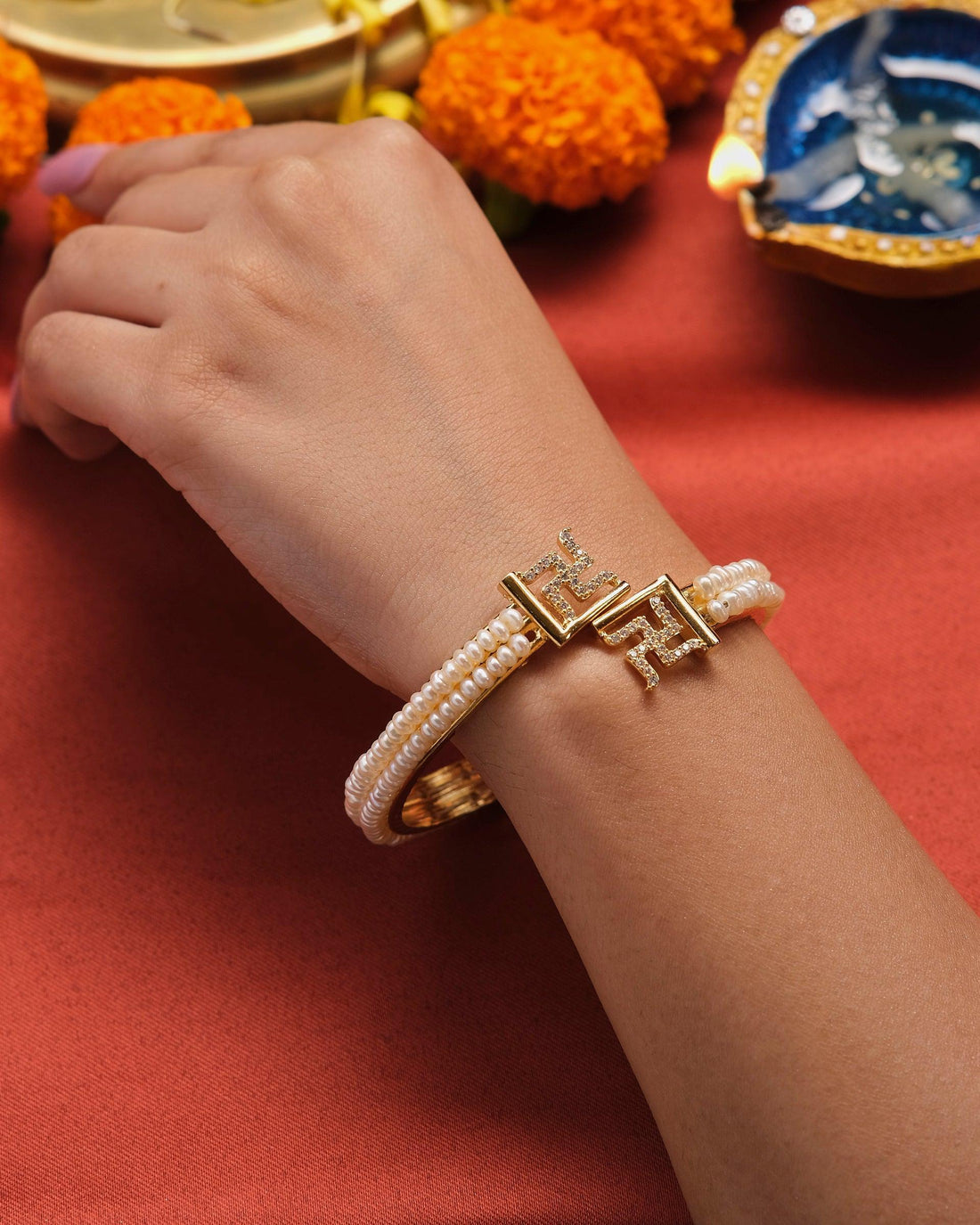 The Shubh Swastik Pearl Bangle by Chandrani Pearls, featuring gold accents and decorative elements, is elegantly displayed on a red box against a blurred background.