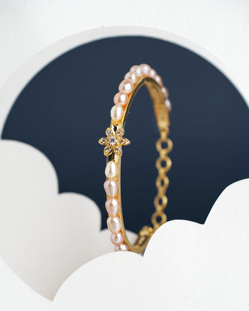 The Astoria Tulip Pearl Bangle by Chandrani Pearls India is a gold bracelet for kids featuring pearls and a floral design, set against a dark backdrop with white cloud-like patterns.