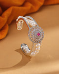 The Gorgeous & Beautiful Shine with Red CZ Bangle by Chandrani Pearls features a floral design with CZ stones, pearls, and a striking red stone, elegantly displayed on beige with orange fabric in the background.
