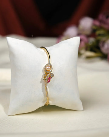 The Joella CZ Stone Bangle by Chandrani Pearls India features an elegant curved design with sparkling CZ stones, pearls, and a pink accent, presented on a white cushion for a glamorous display.