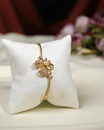 The Kinaya Sparkle CZ Stone Bangle by Chandrani Pearls India features an elegant floral design with pearls and crystals, creating a glamorous look. Presented on a white cushion, the background is softly blurred for emphasis.