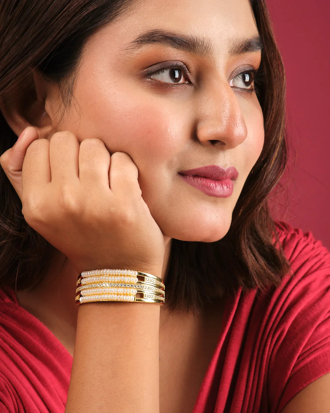 The Natalie CZ Stone Bangle by Chandrani Pearls India features an elegant gold design with pearls and crystals, displayed on a white cushion with blurred purple flowers in the background.