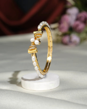 The Spring Design Pearl Bangle by Chandrani Pearls India features gold with pearls and small diamonds, displayed upright on a white round stand against a softly blurred floral background.