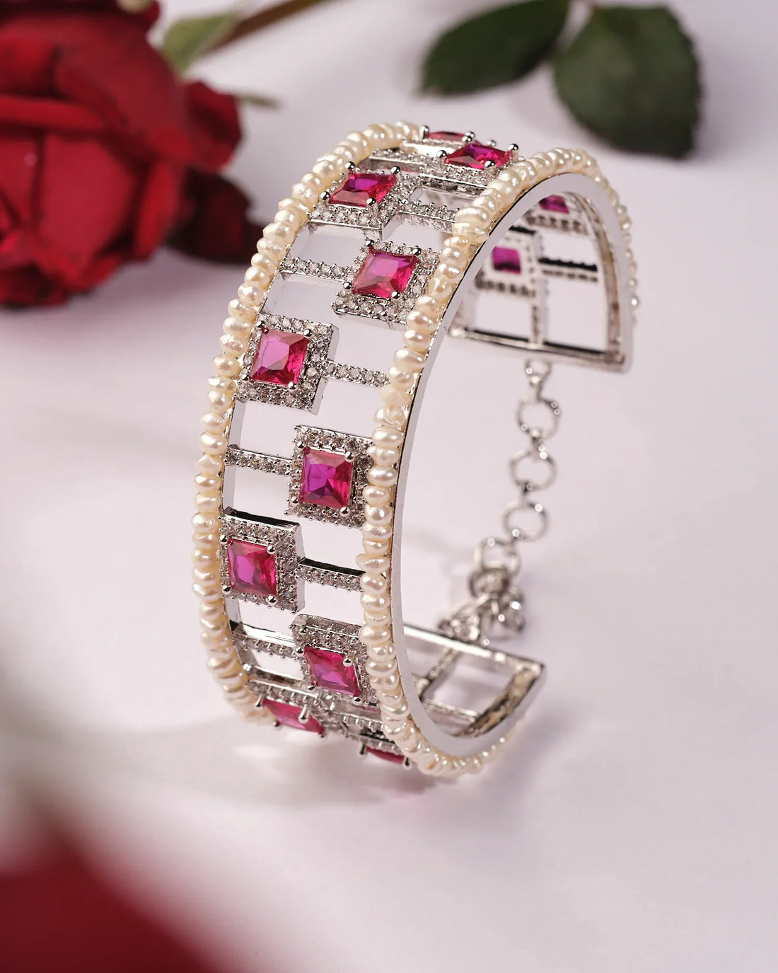 The Kinaya Sparkle CZ Pearl Bangle by Chandrani Pearls India features square red gemstones and luminous pearls, shining luxuriously as it stands upright on a red surface with red roses in the background.