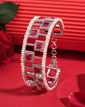 The Kinaya Sparkle CZ Pearl Bangle by Chandrani Pearls India features square red gemstones and luminous pearls, shining luxuriously as it stands upright on a red surface with red roses in the background.