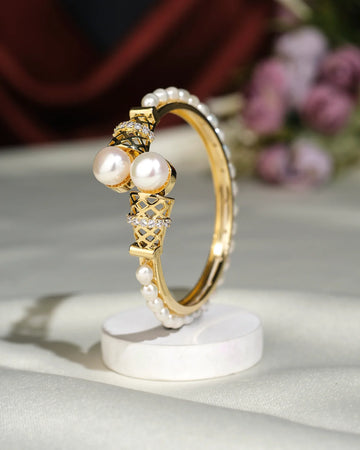 The Trendy White Bangle by Chandrani Pearls India is displayed upright on a white circular stand, set against a blurred floral background.