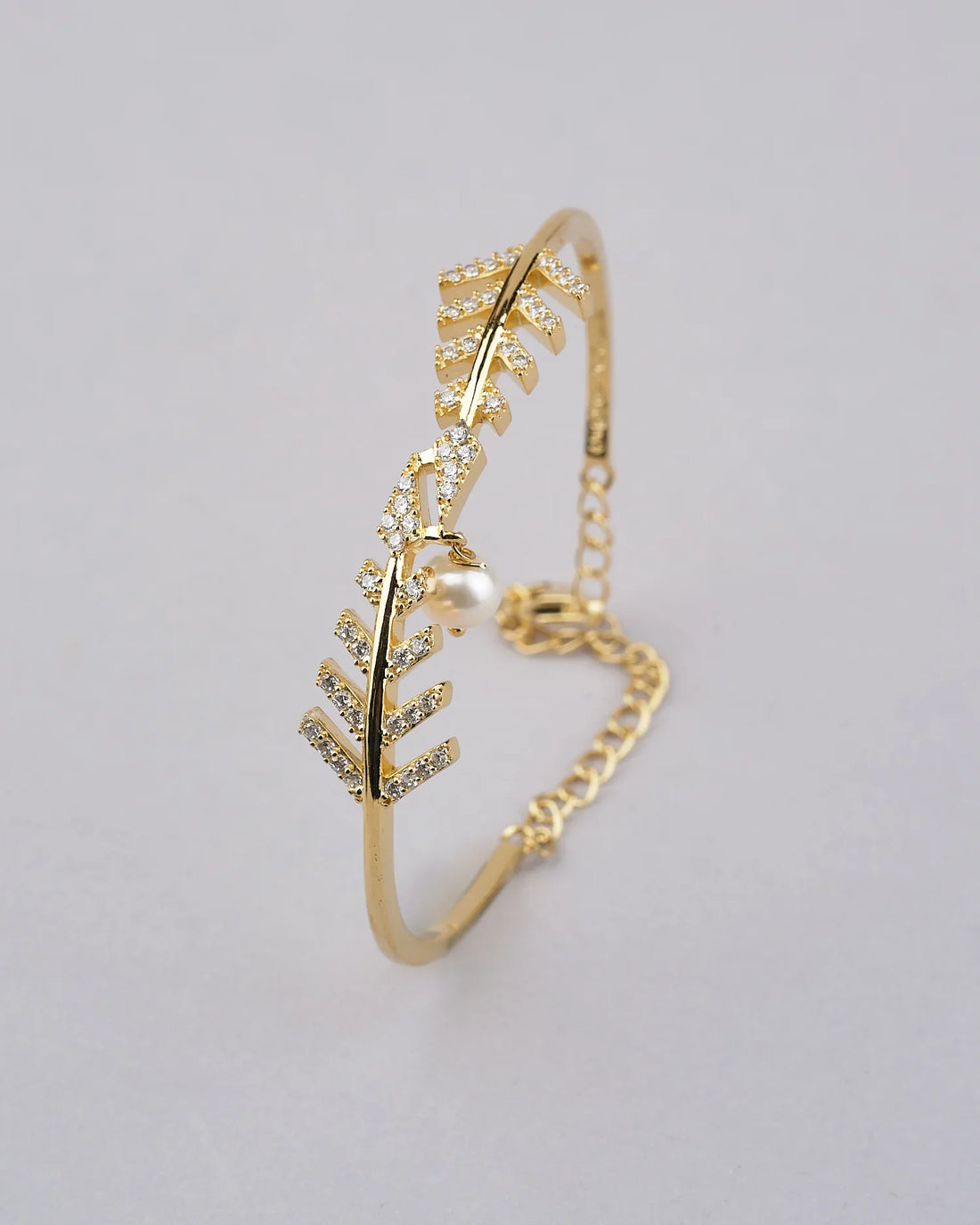 The Ara and Pearl 925 Silver Bangle by Chandrani Pearls India features a leaf motif with crystal accents and a pearl charm, shown on a red surface against a red and pink floral background.