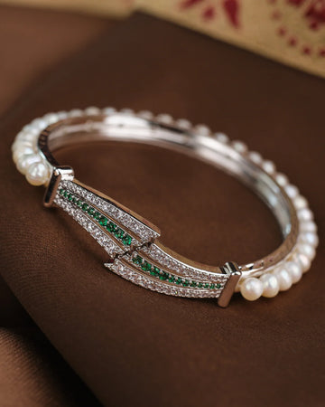 The Starlight Pearl CZ Bangle by Chandrani Pearls, featuring small pearls, green gemstones, and sparkling CZ stones, is elegantly displayed on a brown fabric surface.