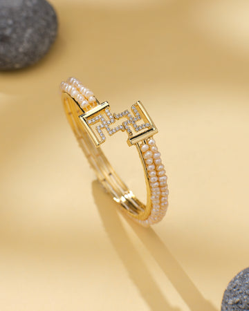 Swasthik White Pearl & CZ Cuff Bangle by Chandrani Pearls—a chic statement piece with two rows of small pearls, CZ stones, and a geometric clasp, styled on a light beige surface with smooth gray stones in the background.