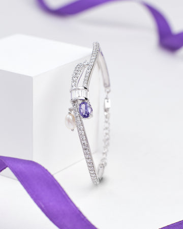 The Elegant Sterling Silver Bangle with Sparkling Cubic Zirconia by Chandrani Pearls features rows of shimmering stones, a teardrop purple gem, a pearl charm, and is displayed with a purple ribbon in the background.
