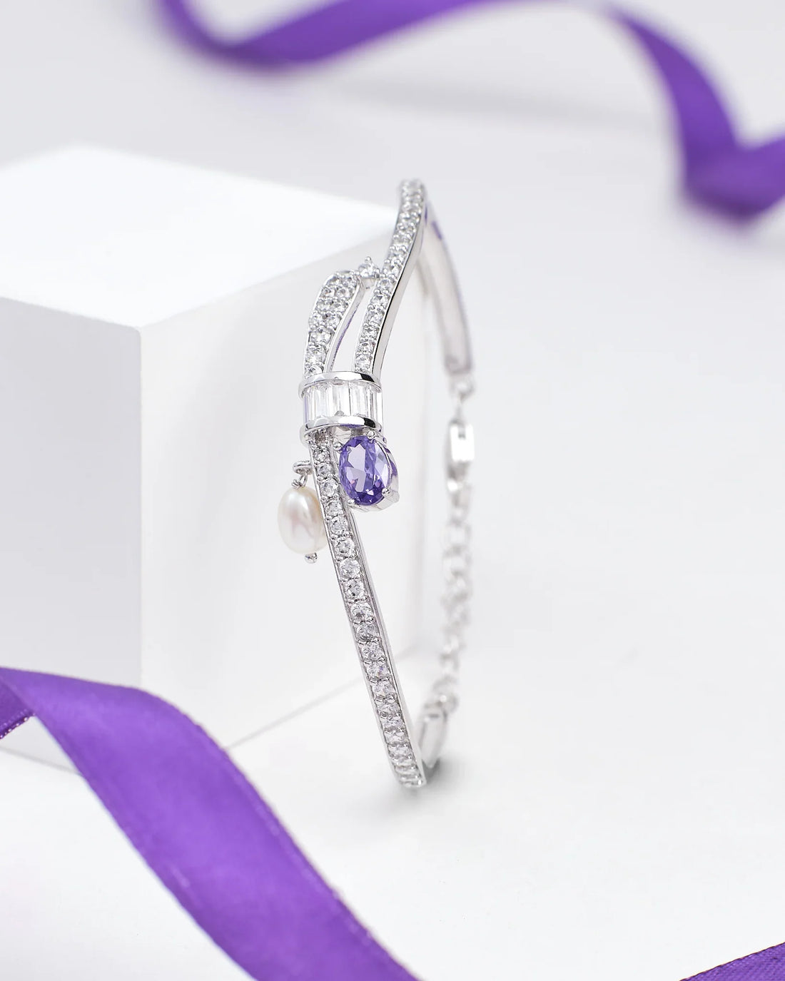 The Elegant Sterling Silver Bangle with Sparkling Cubic Zirconia by Chandrani Pearls features rows of shimmering stones, a teardrop purple gem, a pearl charm, and is displayed with a purple ribbon in the background.
