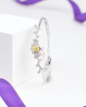 The Chandrani Pearls Everyday Glam 925 Silver Bangle with CZ features a leaf pattern, yellow gemstone, white diamonds, and a pearl charm, beautifully displayed on a white block with purple ribbons in the background.