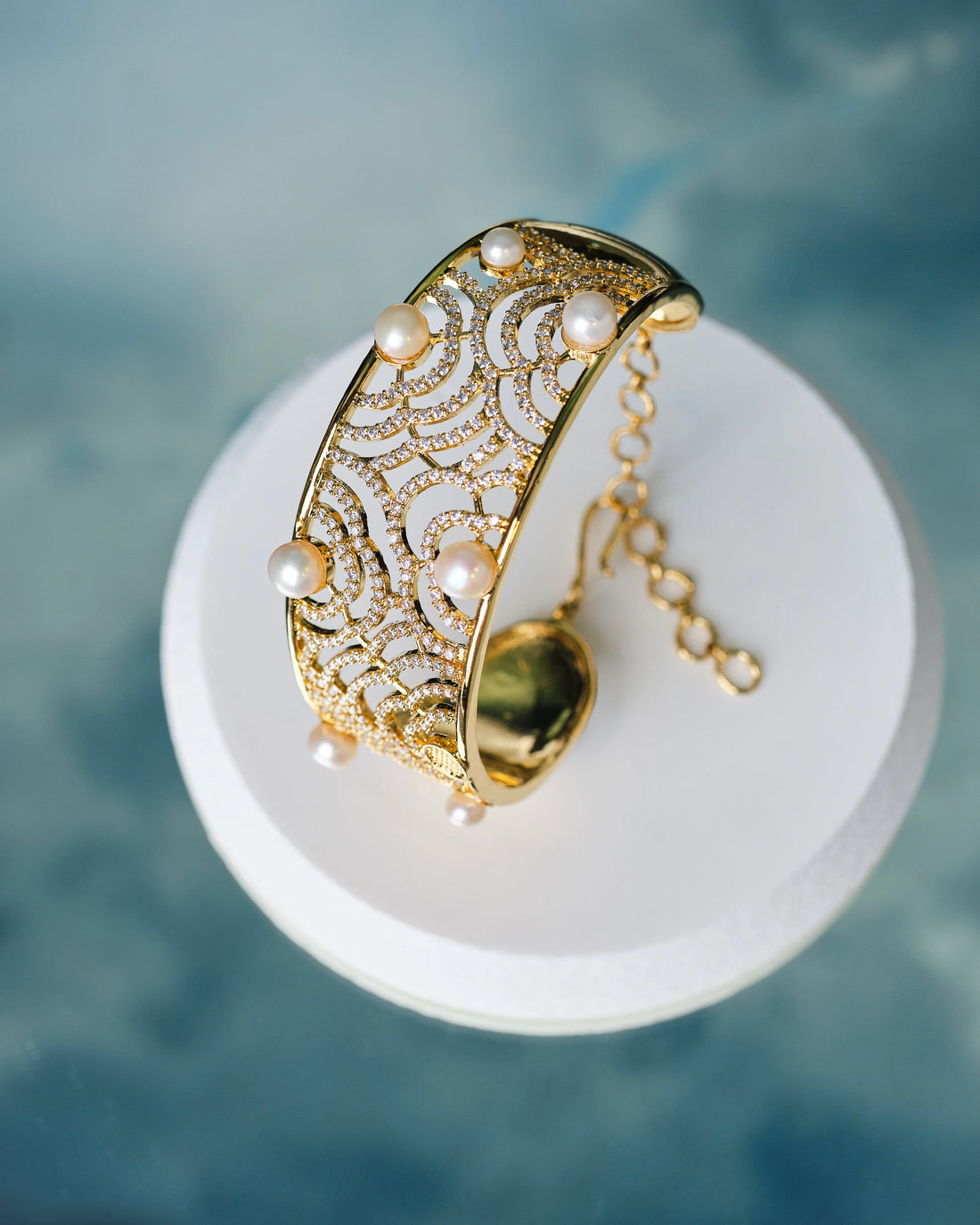 The Stunning pearl bangle accented with sparkling CZ stones by Chandrani Pearls is displayed on a round white platform against a blue background.