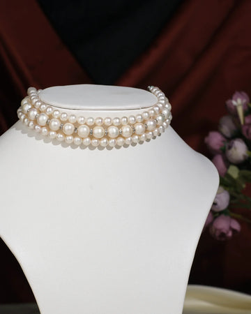 The Elegant Triple Line Choker by Chandrani Pearls India showcases refined glamour on a white jewelry stand, set against a softly blurred floral backdrop.