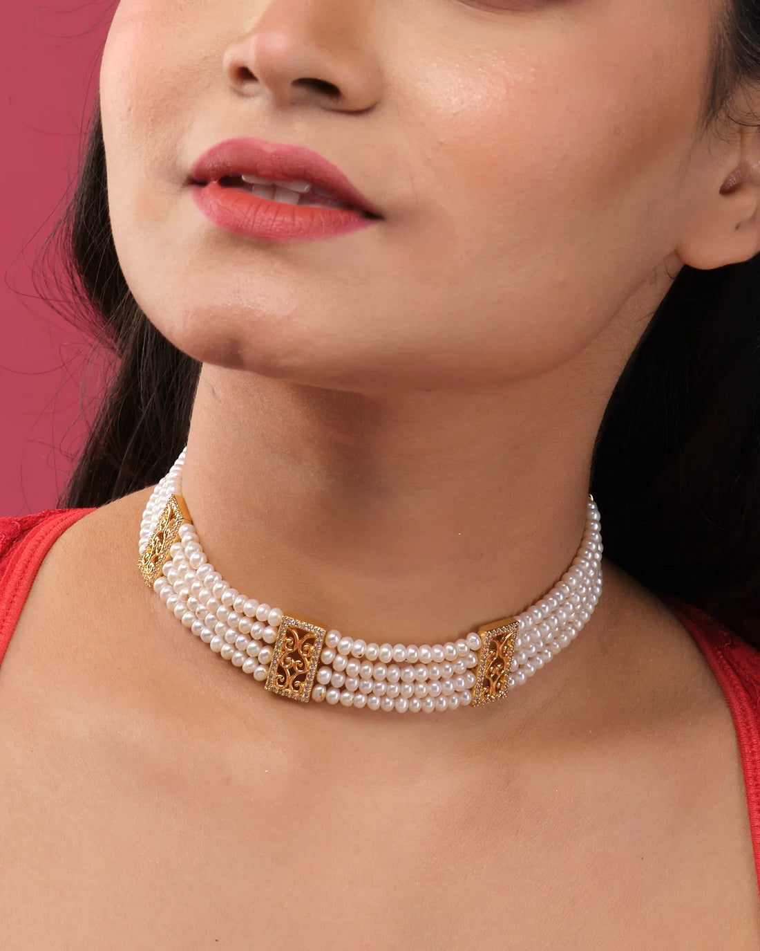 The Beautiful 4 Line Pearl Choker by Chandrani Pearls India features gold rectangular accents, displayed on a white stand against dark draped cloth and a bouquet of purple flowers, exuding luxurious simplicity.