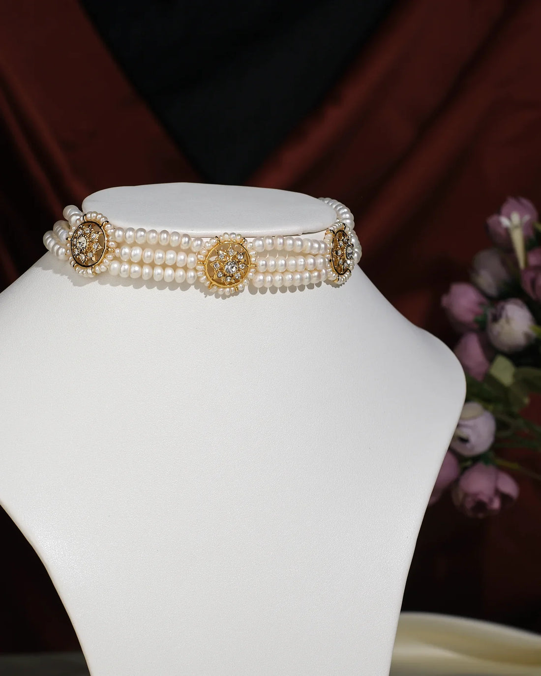 The Trendy 3 Line Pearl Choker by Chandrani Pearls India features gold and rhinestone floral accents, elegantly displayed on a white stand with a blurred flower arrangement in the background.