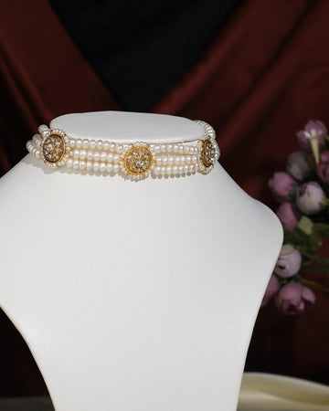 The Trendy 3 Line Pearl Choker by Chandrani Pearls India features gold and rhinestone floral accents, elegantly displayed on a white stand with a blurred flower arrangement in the background.