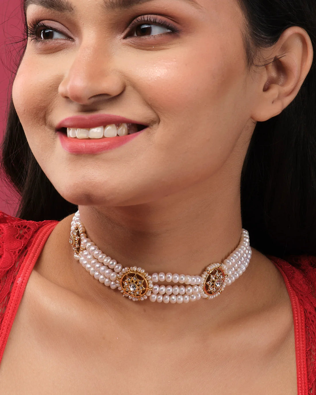 The Trendy 3 Line Pearl Choker by Chandrani Pearls India features gold and rhinestone floral accents, elegantly displayed on a white stand with a blurred flower arrangement in the background.