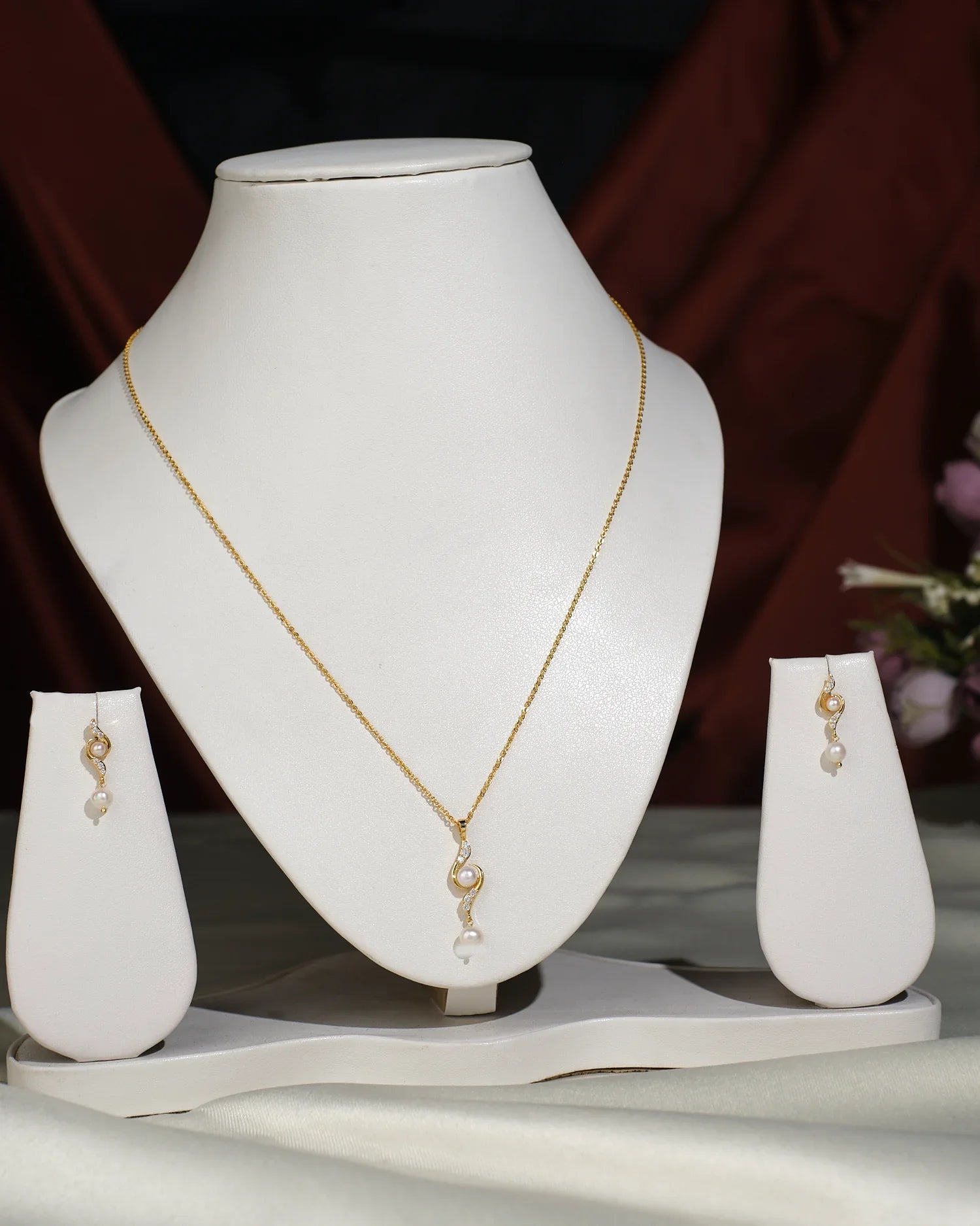 The Pretty Pendant Set by Chandrani Pearls India features a gold pendant and matching earrings with pearl and crystal accents, displayed elegantly on a white stand against a draped background.
