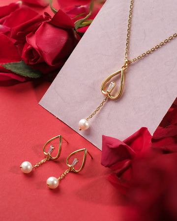 The Heart Silhouette 925 Silver Pendant Set by Chandrani Pearls India features a gold heart design with pearl and crystal accents, paired with matching earrings, displayed on a red surface with roses for timeless elegance.