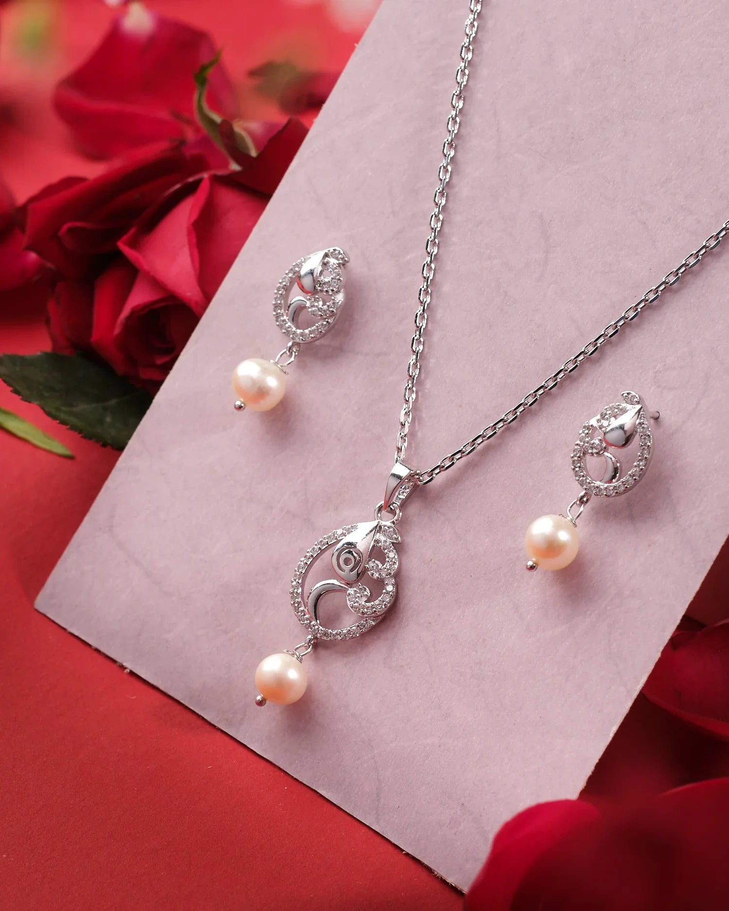 The Marina 925 Silver Pendant Set by Chandrani Pearls India features pearl drops and crystal accents, exuding sophistication. Displayed on a light pink card, it is beautifully surrounded by red roses.