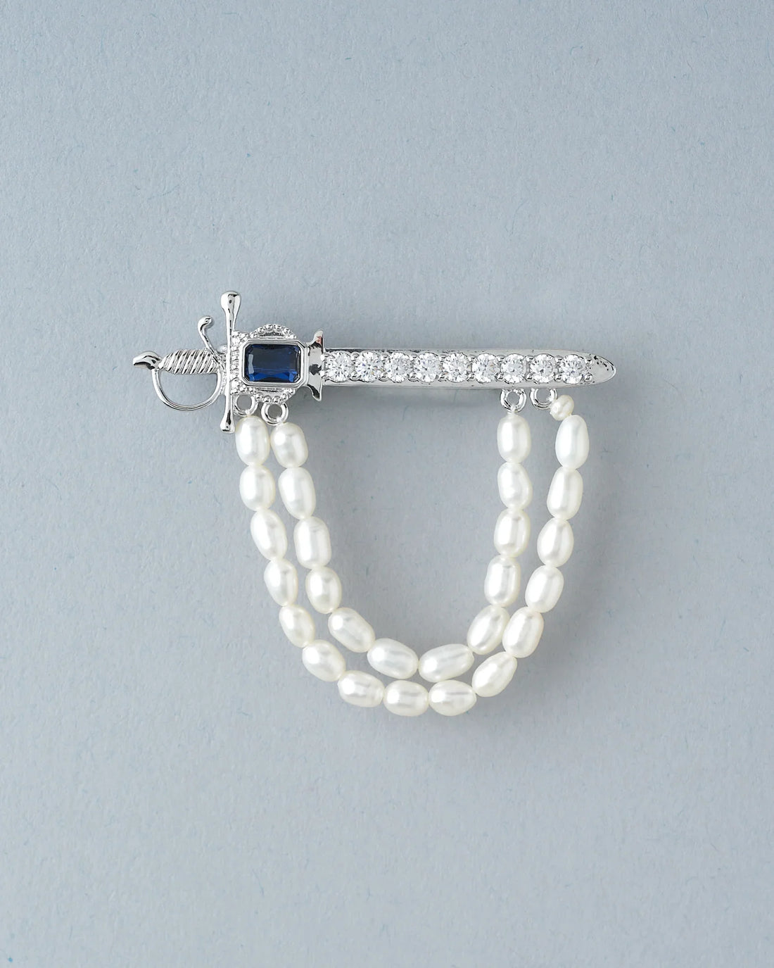 The Husky Gleam Pearl Brooch For Him by Chandrani Pearls, featuring a silver sword design with a black gemstone, rhinestones, and double pearl strands, exudes elegance atop a black surface with rust-colored fabric.