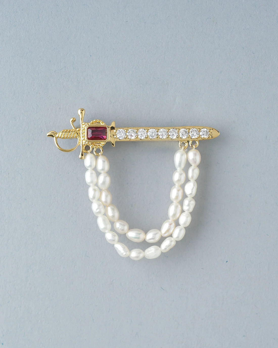 The Brawny Waves Pearl Brooch For Him by Chandrani Pearls features a gold sword shape, red gemstone, clear stones, and two strands of white pearls—an exquisite piece displayed on a dark surface with rust-colored fabric in the background.