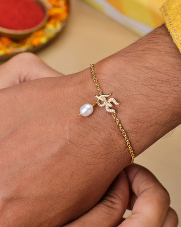 The Trishul Kavacha Pearl Bracelet by Chandrani Pearls, featuring a gold chain, pearl charm, and rhinestone Om symbol, is worn to symbolize spiritual strength and devotion to Lord Shiva.
