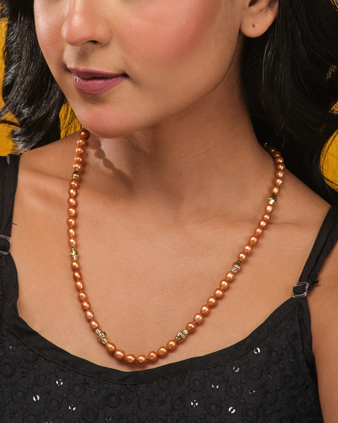 The Aurora Pearl Necklace by Chandrani Pearls, featuring brown pearls and gold accents, is showcased on a tan surface with orange textured fabric in the background.