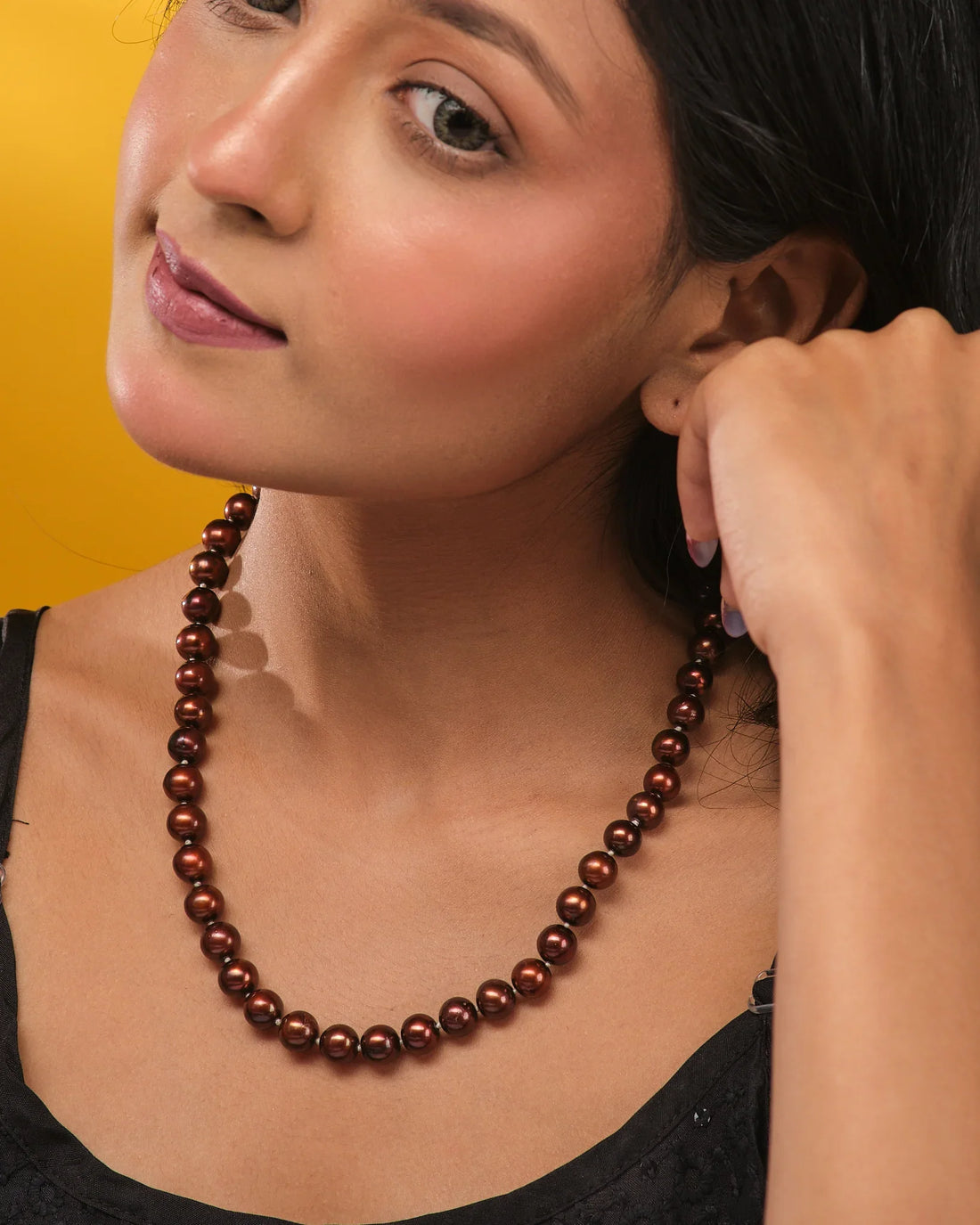 The Luna Pearl Round Necklace by Chandrani Pearls, featuring dark brown pearls and a gold clasp, rests on a tan surface beside orange textured fabric.