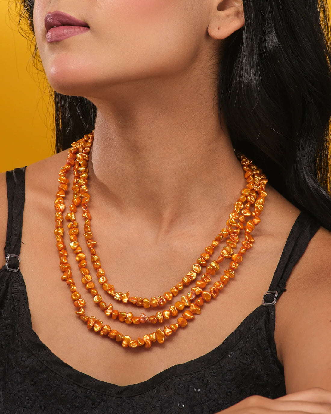 The Celeste Un-Shaped Gold Layered Necklace by Chandrani Pearls features three strands of irregular gold-colored beads with a clasp, displayed on a light brown surface beside orange textured fabric—a trendy choice for jewelry lovers.