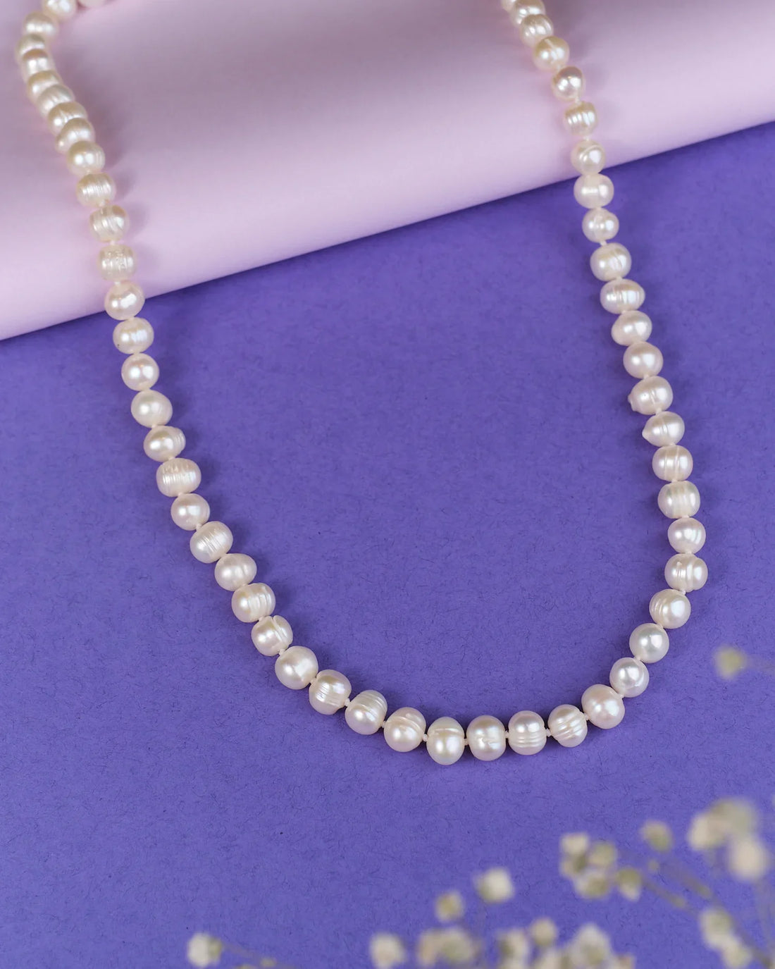 The Heavenly Pearl Necklace by Chandrani Pearls features elegant white pearls arranged in a circular form on a purple and pink background with delicate white flowers in the foreground.