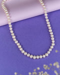 The Heavenly Pearl Necklace by Chandrani Pearls features elegant white pearls arranged in a circular form on a purple and pink background with delicate white flowers in the foreground.
