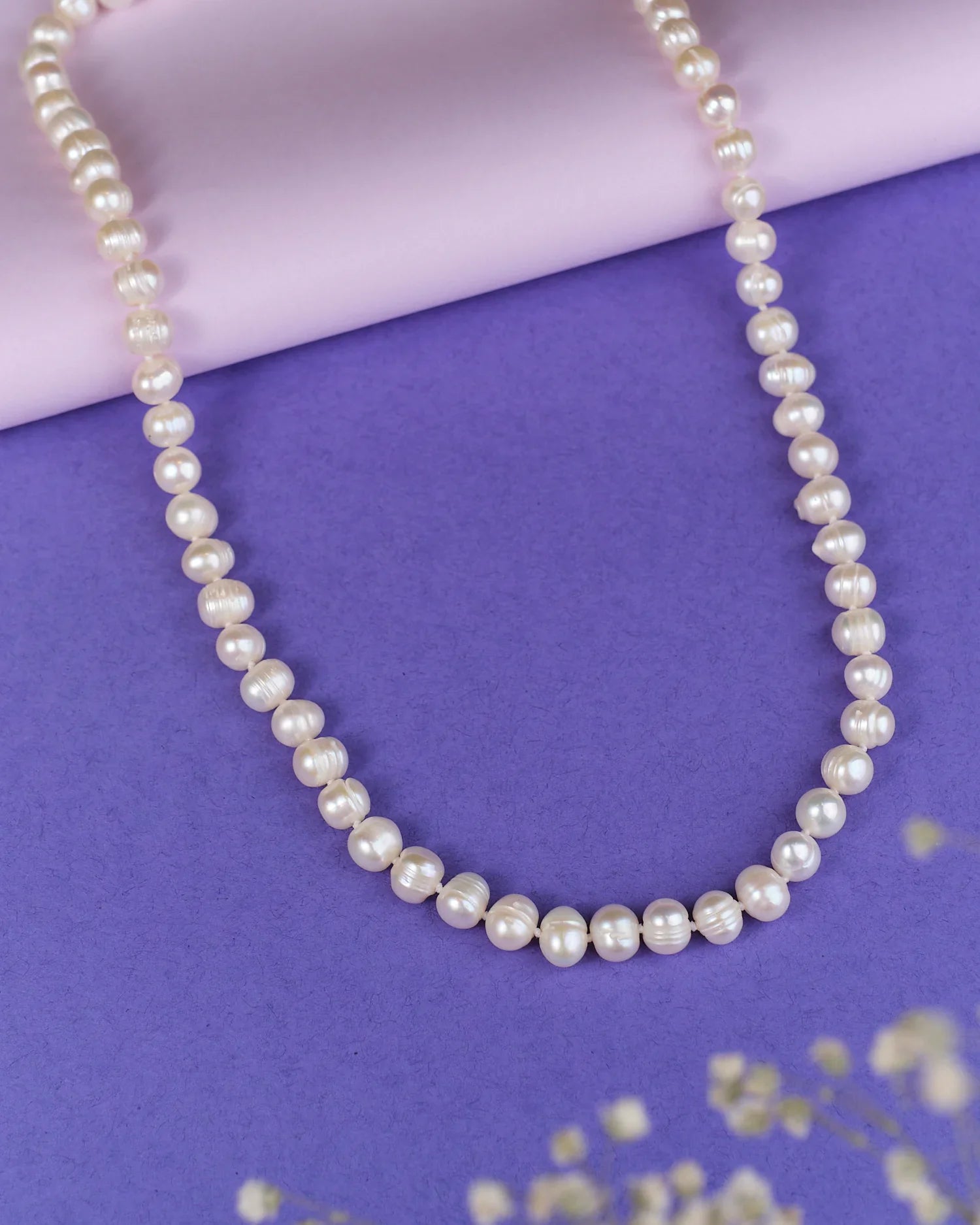 The Heavenly Pearl Necklace by Chandrani Pearls features elegant white pearls arranged in a circular form on a purple and pink background with delicate white flowers in the foreground.
