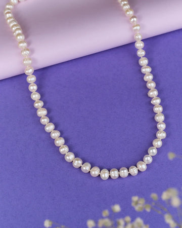 The Heavenly Pearl Necklace by Chandrani Pearls features elegant white pearls arranged in a circular form on a purple and pink background with delicate white flowers in the foreground.
