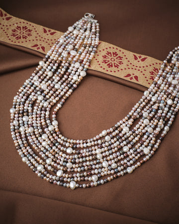 The Chandrani Pearls Fashion Multicolor 10 Line Pearl Mala, featuring white, pink, and lavender pearls, is displayed on brown fabric with a decorative ribbon—an elegant choice for wedding jewelry.