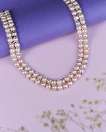 The Lavana-The Coast Memoirs Pearl Necklace by Chandrani Pearls, a luxurious double-stranded piece with pink and white pearls, gracefully drapes over a purple cylinder on a pink surface. Adorned with small white flowers, it is the perfect versatile accessory for any ensemble.