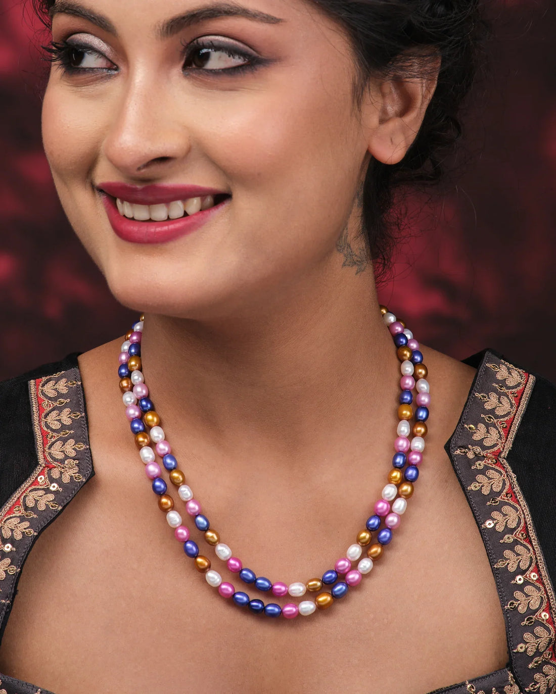 The Multi Colour Pearl Necklace by Chandrani Pearls features blue, pink, purple, and gold pearls arranged on a pink surface beside a folded purple cloth with white leaf patterns.