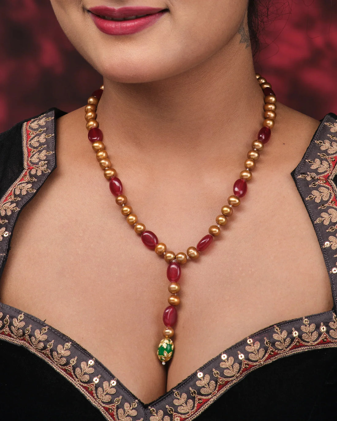 The Elegant Beads & Pearl Mala by Chandrani Pearls—featuring red, gold, and green beads with iridescent pearls—is displayed on a pink surface beside purple fabric adorned with white leaf motifs.