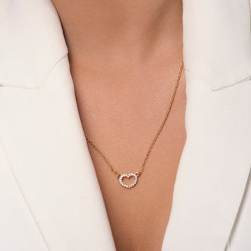 A person wearing a white blazer and the Guardian Heart Pendant by Chandrani Pearls, a gold heart necklace adorned with sparkling cubic zirconia stones.