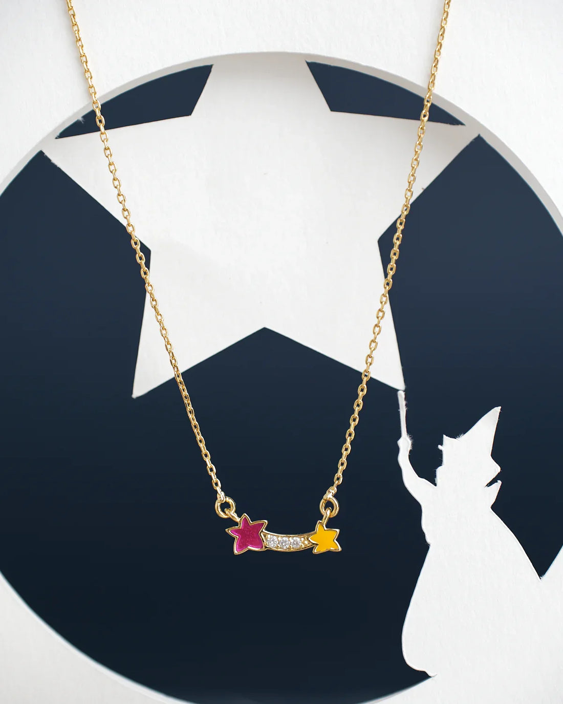 The Dual Star Beautiful Pendant by Chandrani Pearls India is a gold necklace featuring two central stars—one pink, one yellow—linked by clear stones, with a silhouette of a figure holding a wand with a star cutout in the background.
