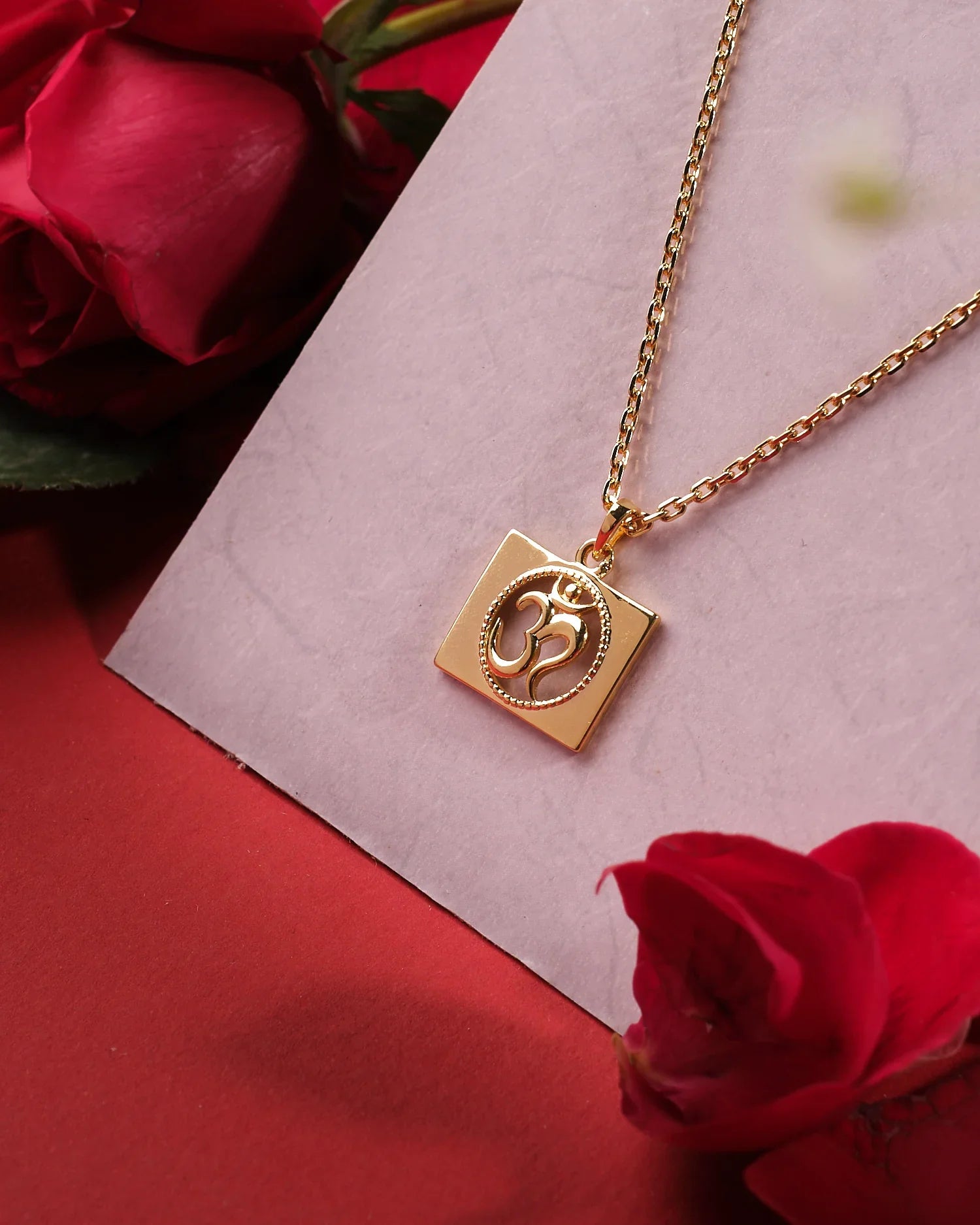 The Omkara 925 Silver Pendant by Chandrani Pearls India features an Om symbol on a square charm, elegantly displayed on a light pink surface with red roses for a refined touch.