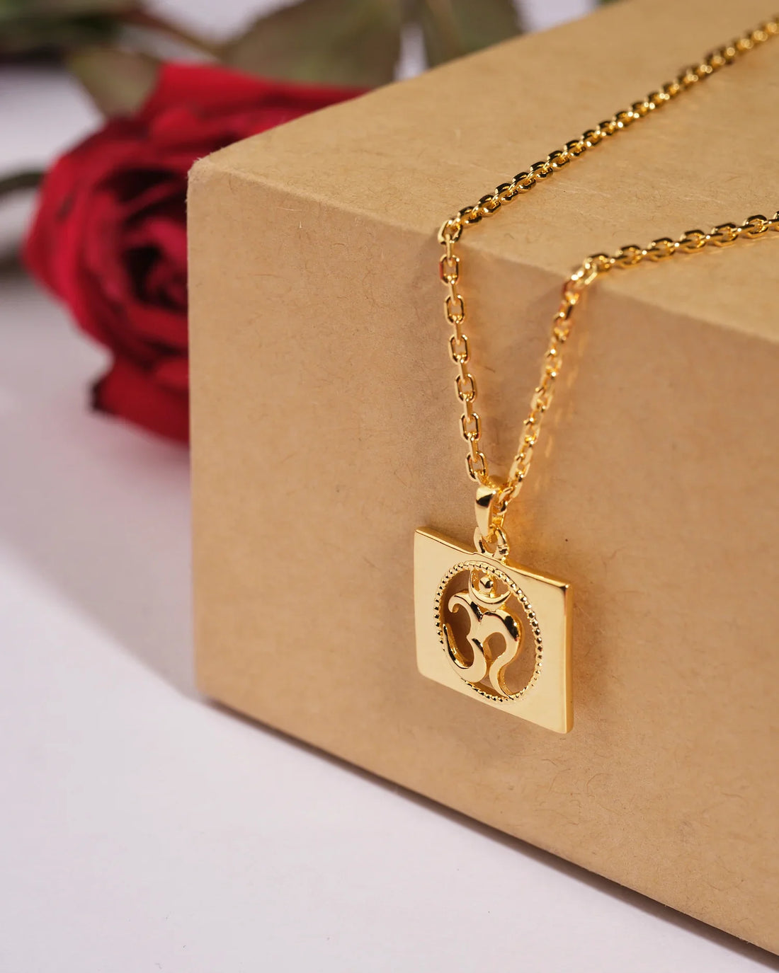 The Omkara 925 Silver Pendant by Chandrani Pearls India features an Om symbol on a square charm, elegantly displayed on a light pink surface with red roses for a refined touch.