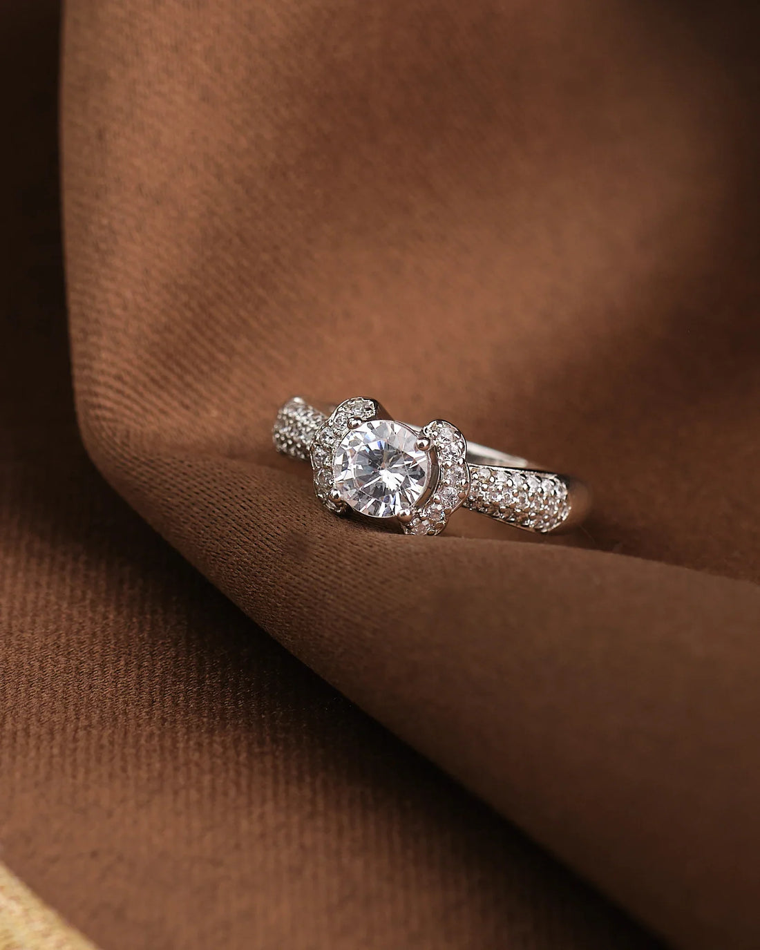 The Solitaire Silver Ring Sleek and Modern by Chandrani Pearls features a large round central gemstone and a band of smaller stones, elegantly showcased on a brown fabric background.
