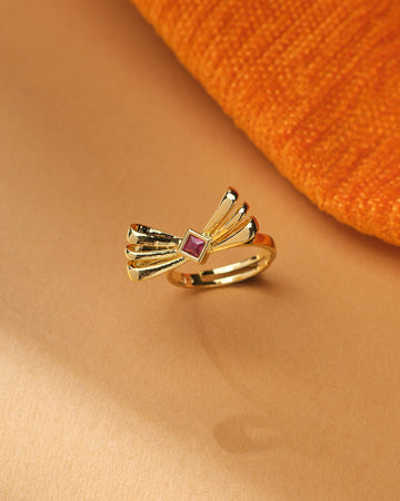 The Trendy Beautiful Ring by Chandrani Pearls, featuring a gold polish, fan-shaped bow design, and central square CZ stone, is displayed on a beige surface beside orange textured fabric.