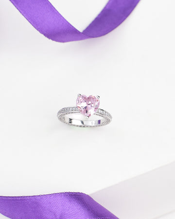 The Heart Inspired Demifine Silver Ring with Pink Sparkling CZ by Chandrani Pearls is a romantic jewelry piece, featuring a heart shape and shimmering CZ, displayed on a white surface with a purple ribbon—ideal for any occasion.