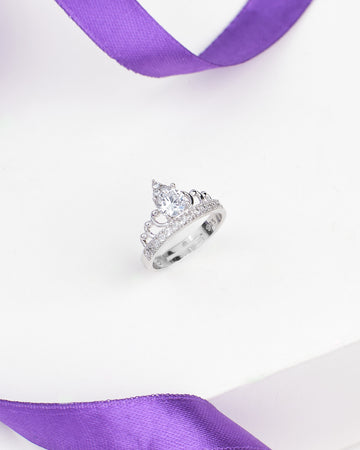 The Romantic Demifine Silver Ring with Solitaire CZ Stone by Chandrani Pearls is shown on a white surface with a purple ribbon, highlighting its elegant tiara-inspired design.